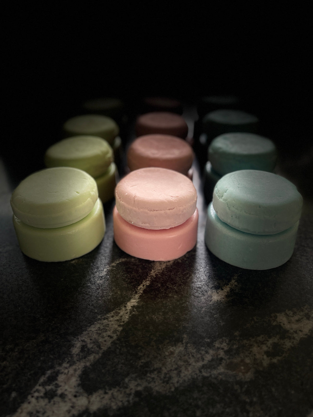 Three rows of green, pink, and blue conditioner and shampoo bars layered on each other, sitting on a dark marbled surface