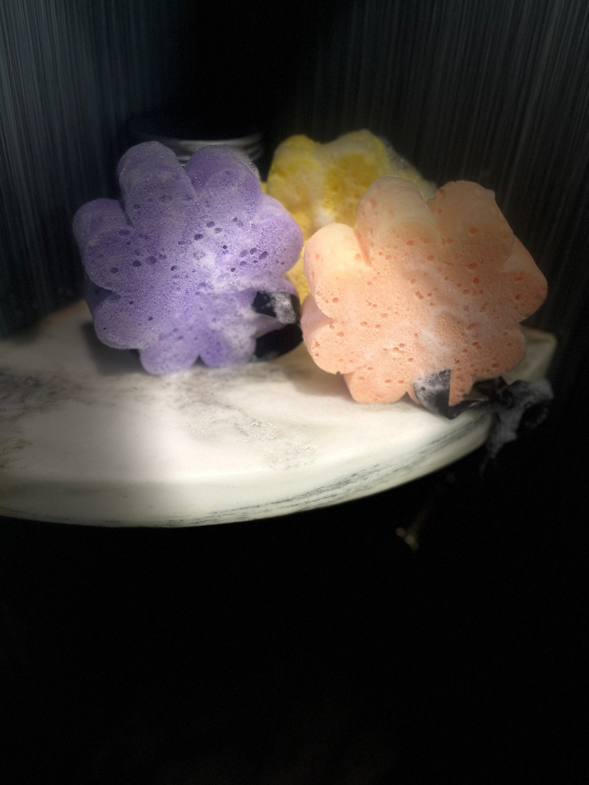 Three flower-shaped soap sponges, one purple and one peach, resting on a white marble surface with a yellow sponge partially visible behind them.