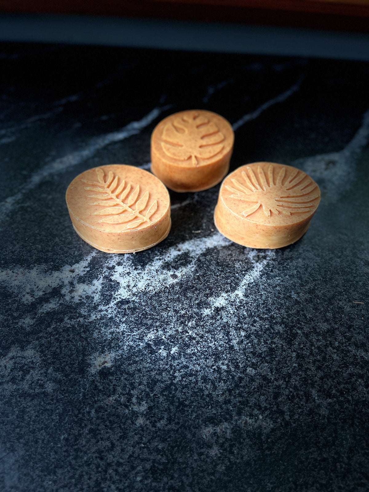 Three oval face soaps with embossed tropical leaf designs, arranged on a dark marbled countertop. 