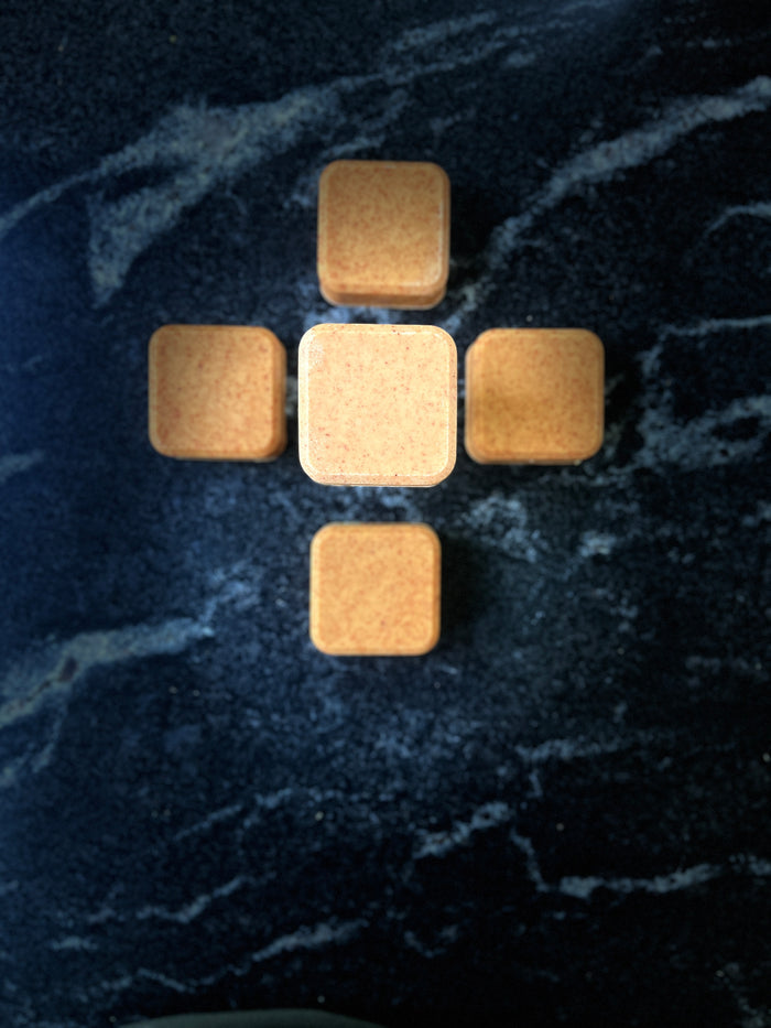 Five small square face soap arranged in a cross pattern on a dark marbled surface.