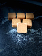 Six small square face soap arranged on a dark textured surface, with one in front in sharper focus.