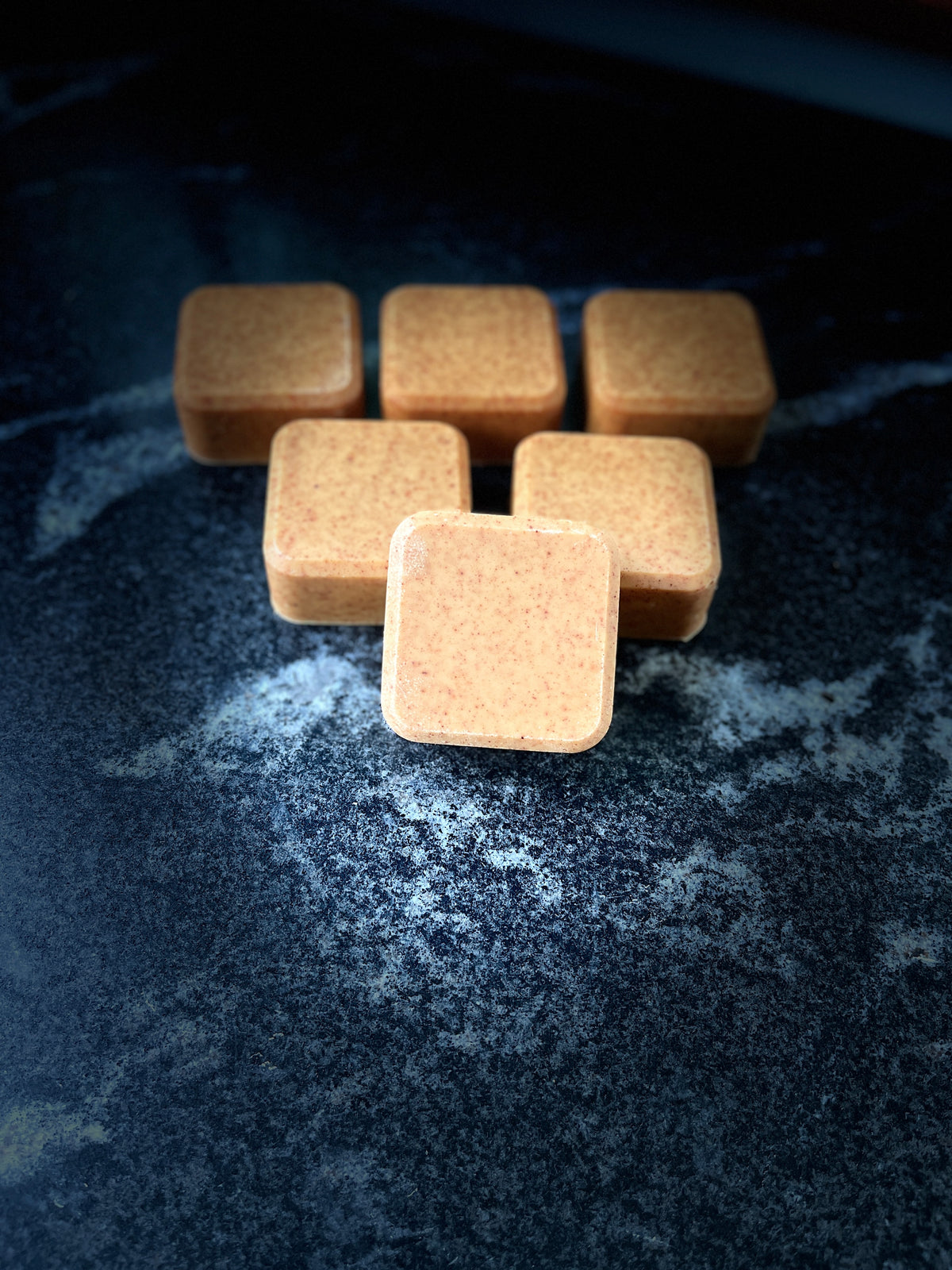 Six small square face soap arranged on a dark textured surface, with one in front in sharper focus.