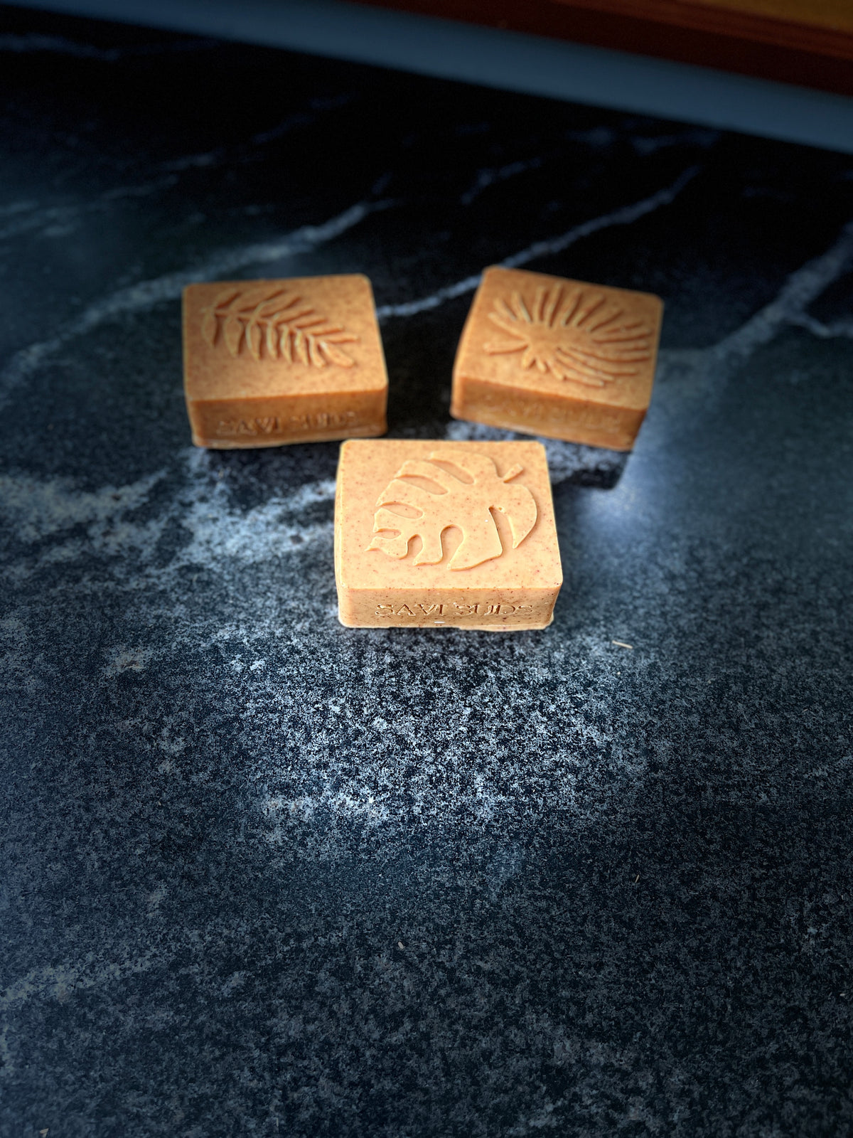 Three square face soaps with embossed tropical leaf designs, arranged on a dark marbled countertop. The front block is upright and shows the brand name 'SAVI SUDS' stamped on the side.