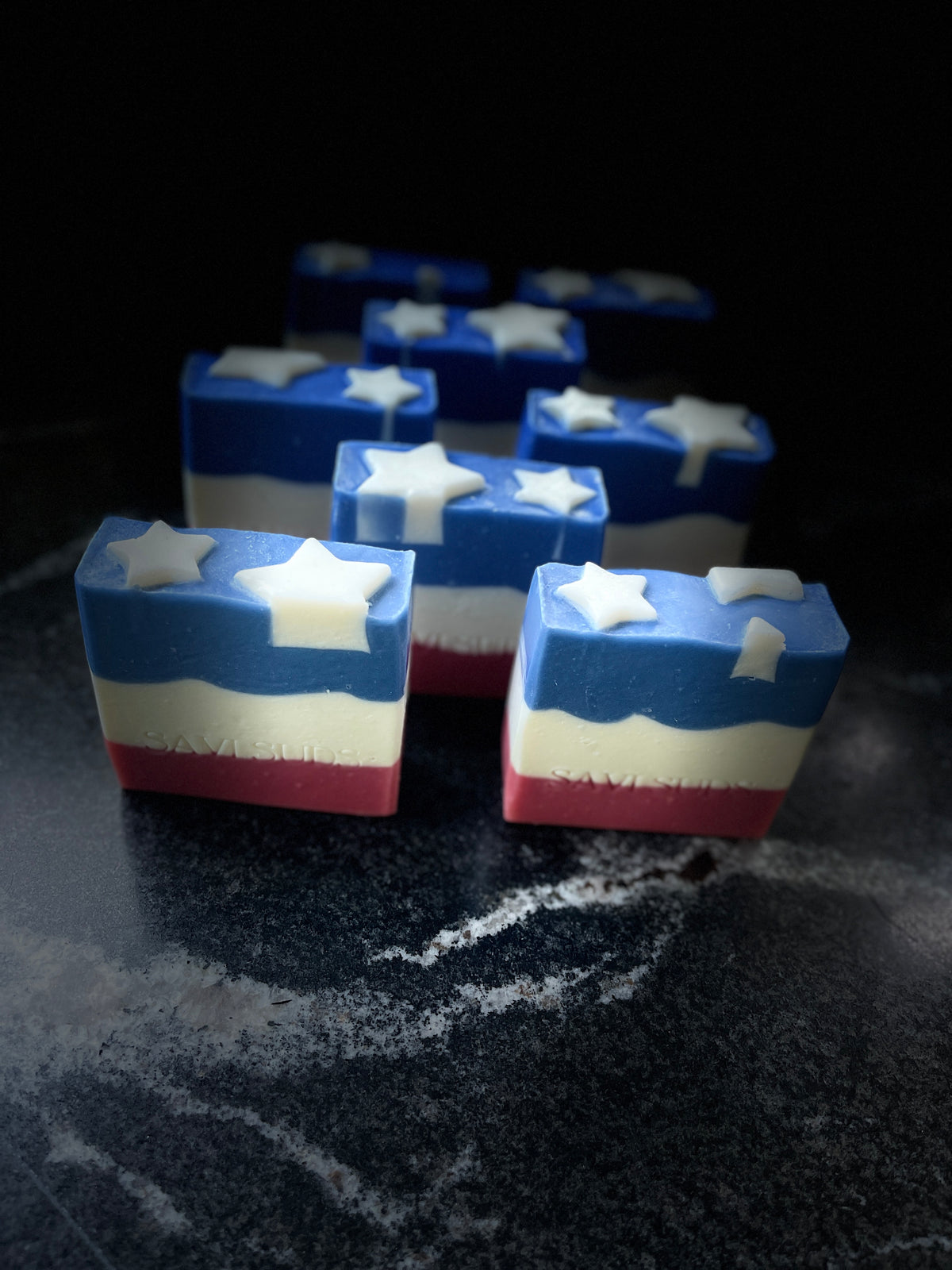 Several square layered bars with a blue top, white star detail, and red-white-blue horizontal layers, arranged on a dark marble-like surface against a black background.