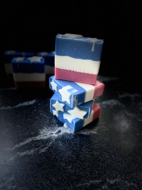 Three handmade square bars of soap stacked on a dark surface, decorated with a red, white, and blue American flag theme including white stars, with more soap bars blurred in the background.
