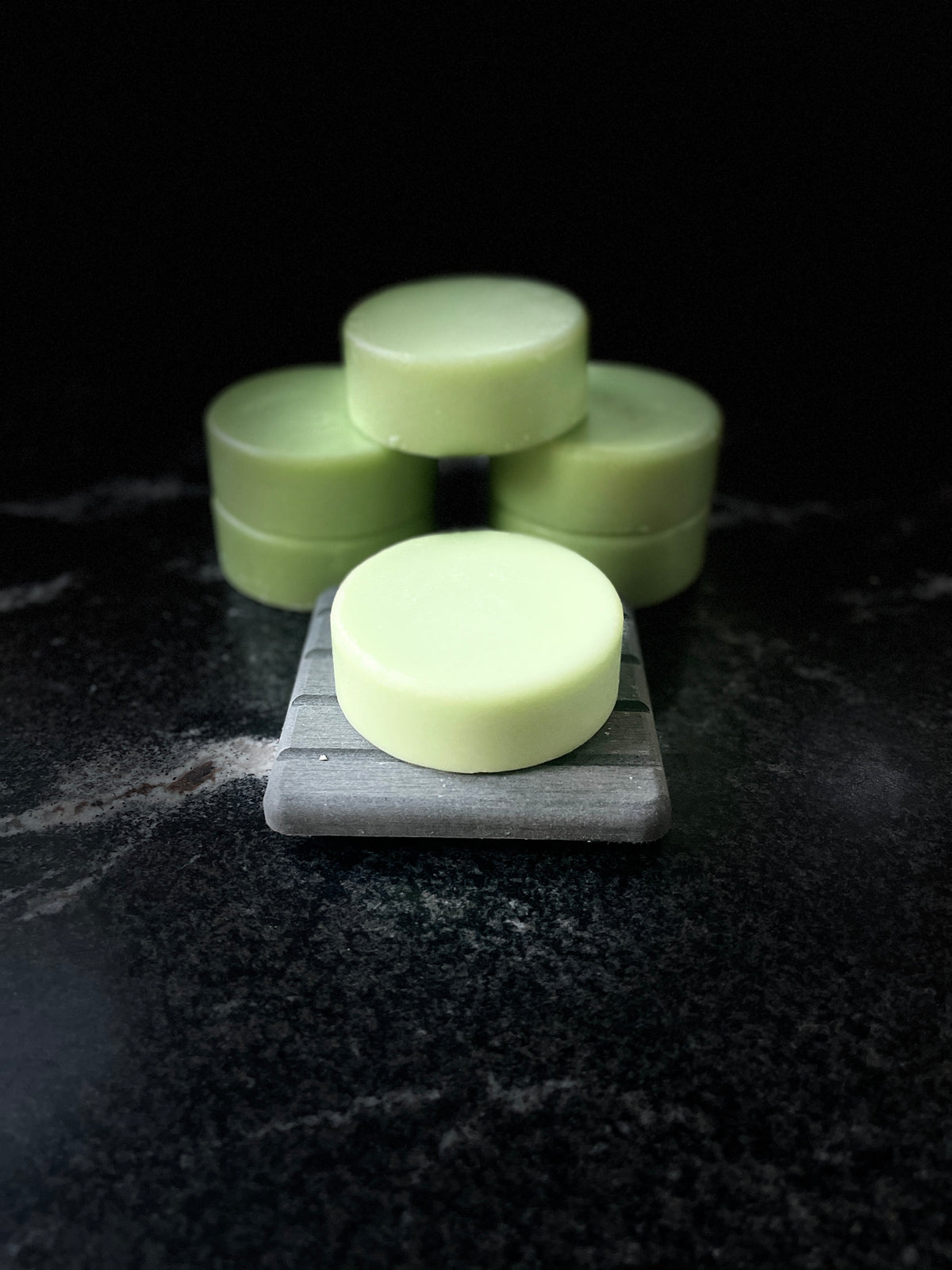 Pale green round Baja Breeze conditioner bars arranged on a dark marble surface, with one bar in the foreground resting on a gray square block and the others stacked behind it against a black background.