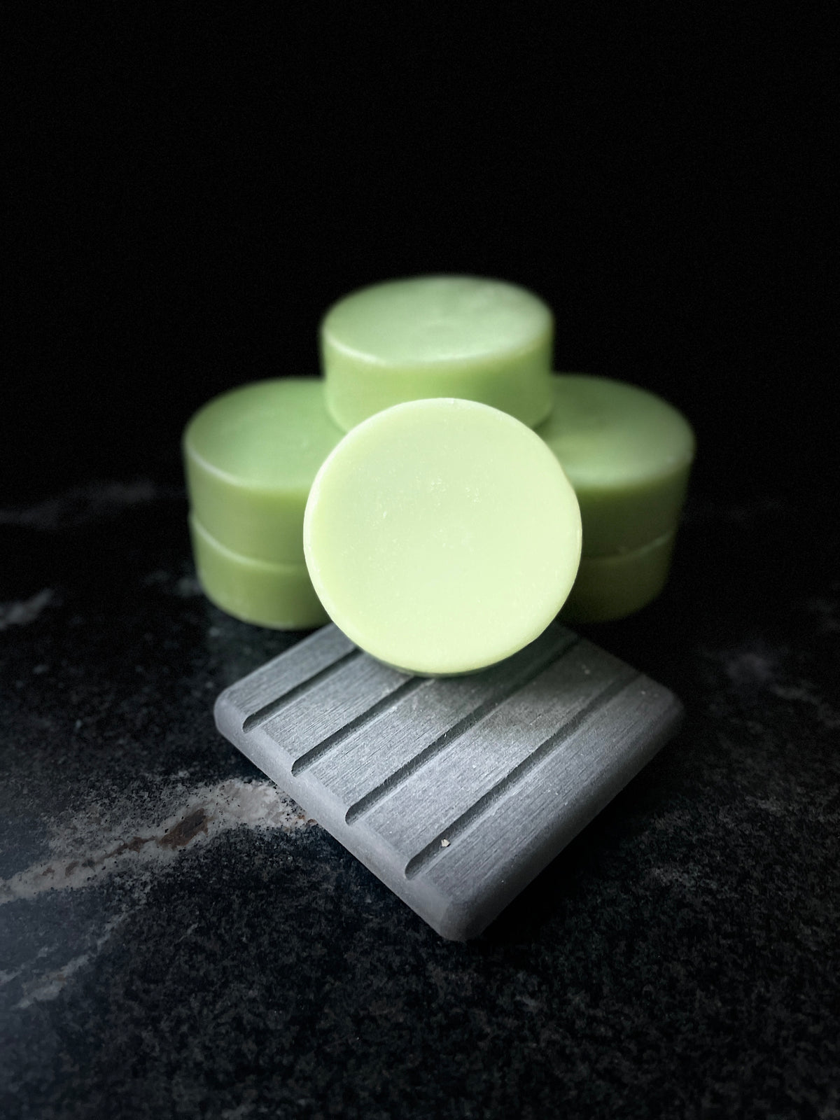 Pale green round Baja Breeze Conditioner bars arranged with one in the foreground resting on a gray square dish, set against a dark black background.