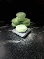 A stack of pale green solid shampoo bars, with one in the foreground resting on a small gray rectangular dish, set against a dark background