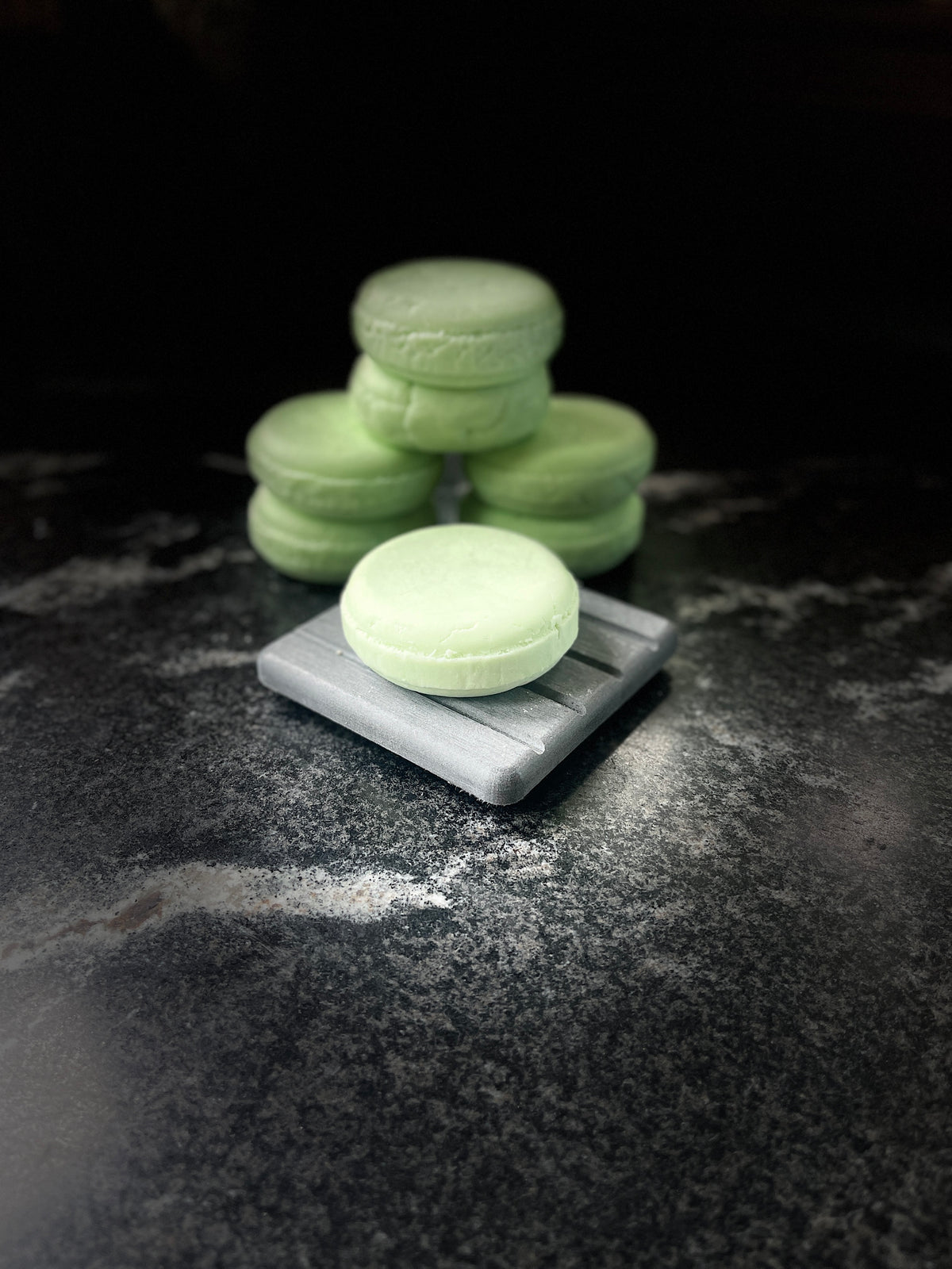 A stack of pale green solid shampoo bars, with one in the foreground resting on a small gray rectangular dish, set against a dark background