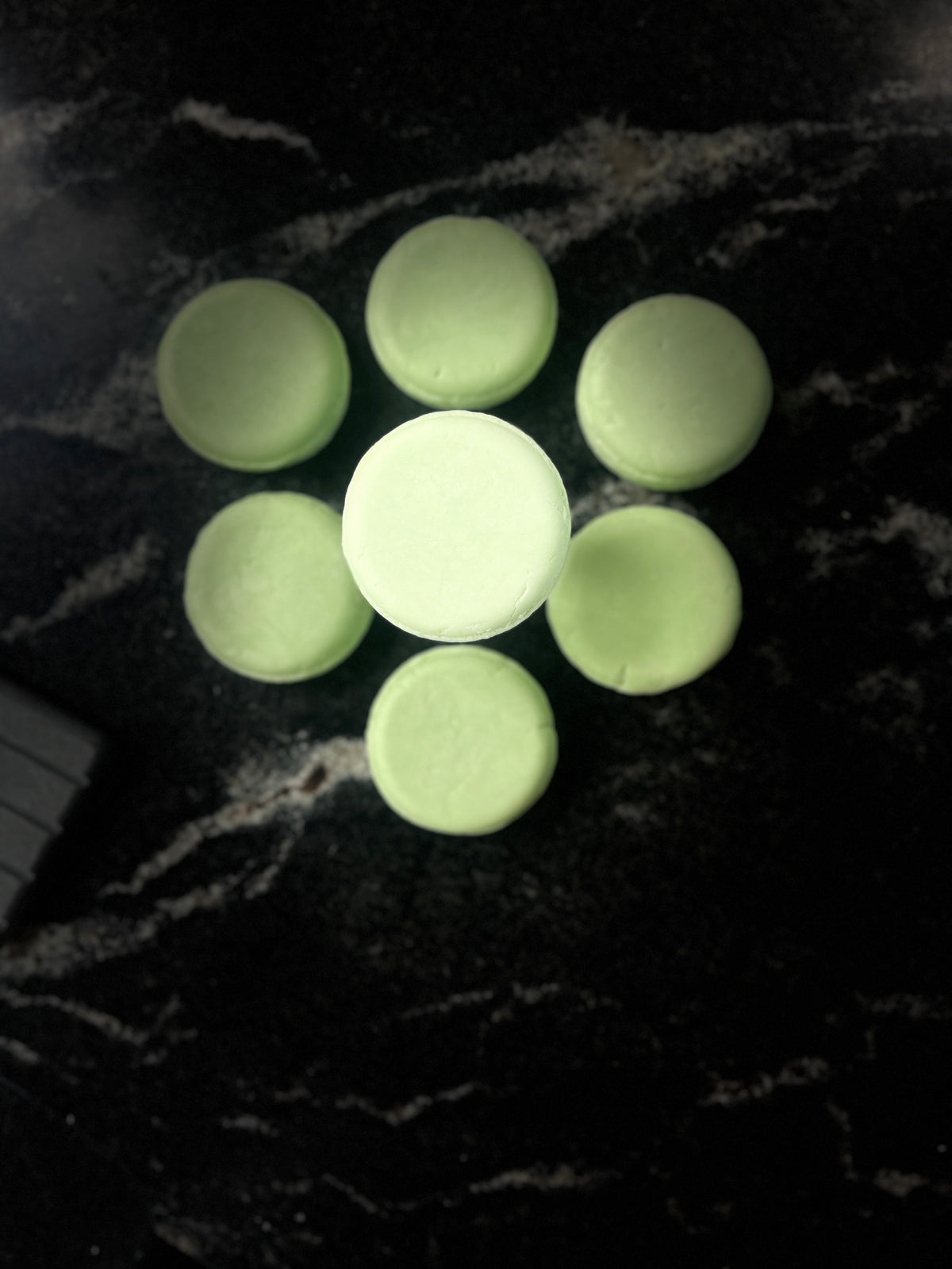 Six pale green shampoo bars arranged in a flower-like pattern on a dark marbled surface, with one item centered and facing forward.