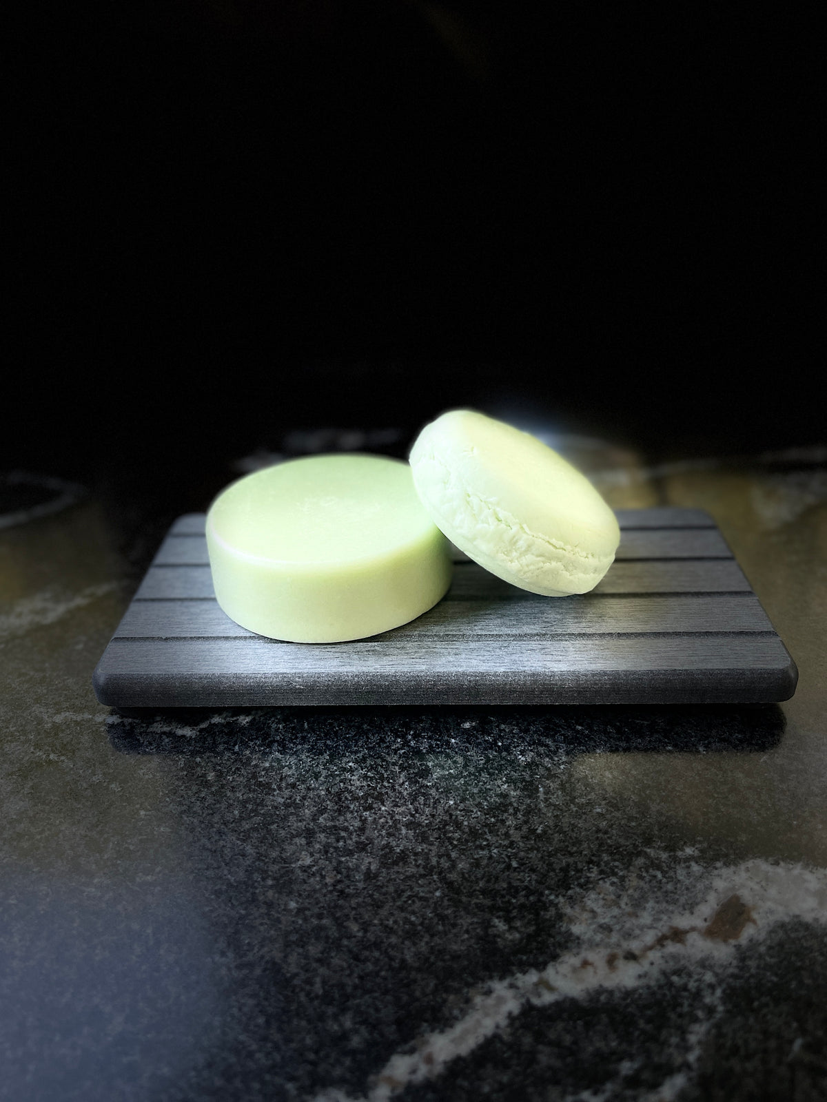 Two pale green shampoo and conditioner bars resting on a dark gray rectangular tray against a black background.