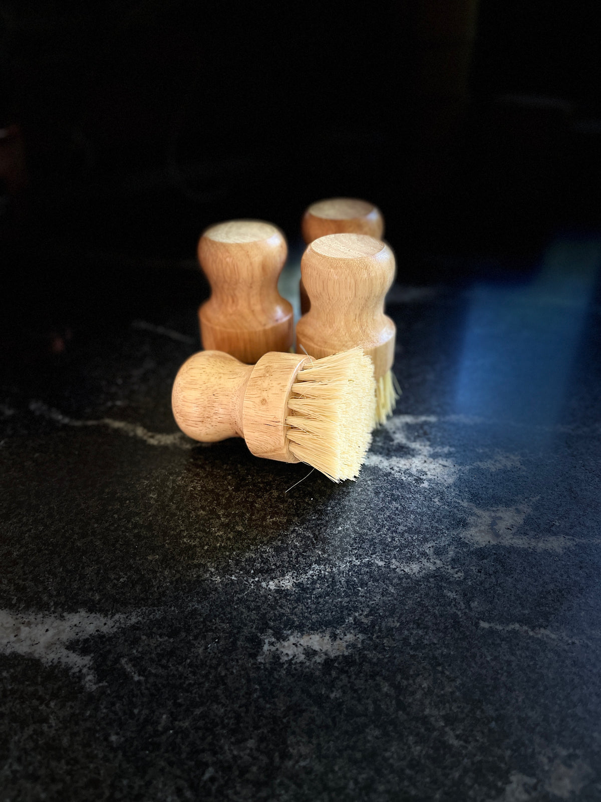 Four small wooden-handled cleaning brushes with light natural bristles arranged on a dark stone surface.