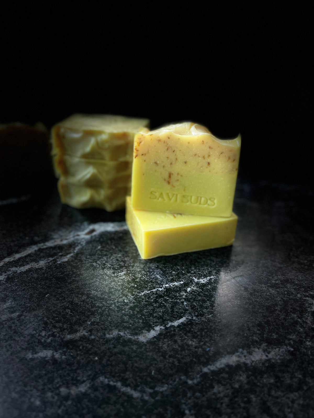 Two square yellow handmade soap bars with botanical inclusions, one stacked on top of the other and stamped with the brand name 'SAVI SUDS', set on a dark marble surface against a black background with more bars blurred in the background