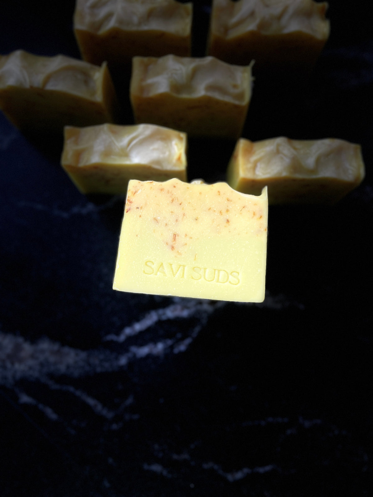 A handmade soap bar in the foreground with 'SAVISUDS' stamped on it, surrounded by several other soap bars with a dark marbled background