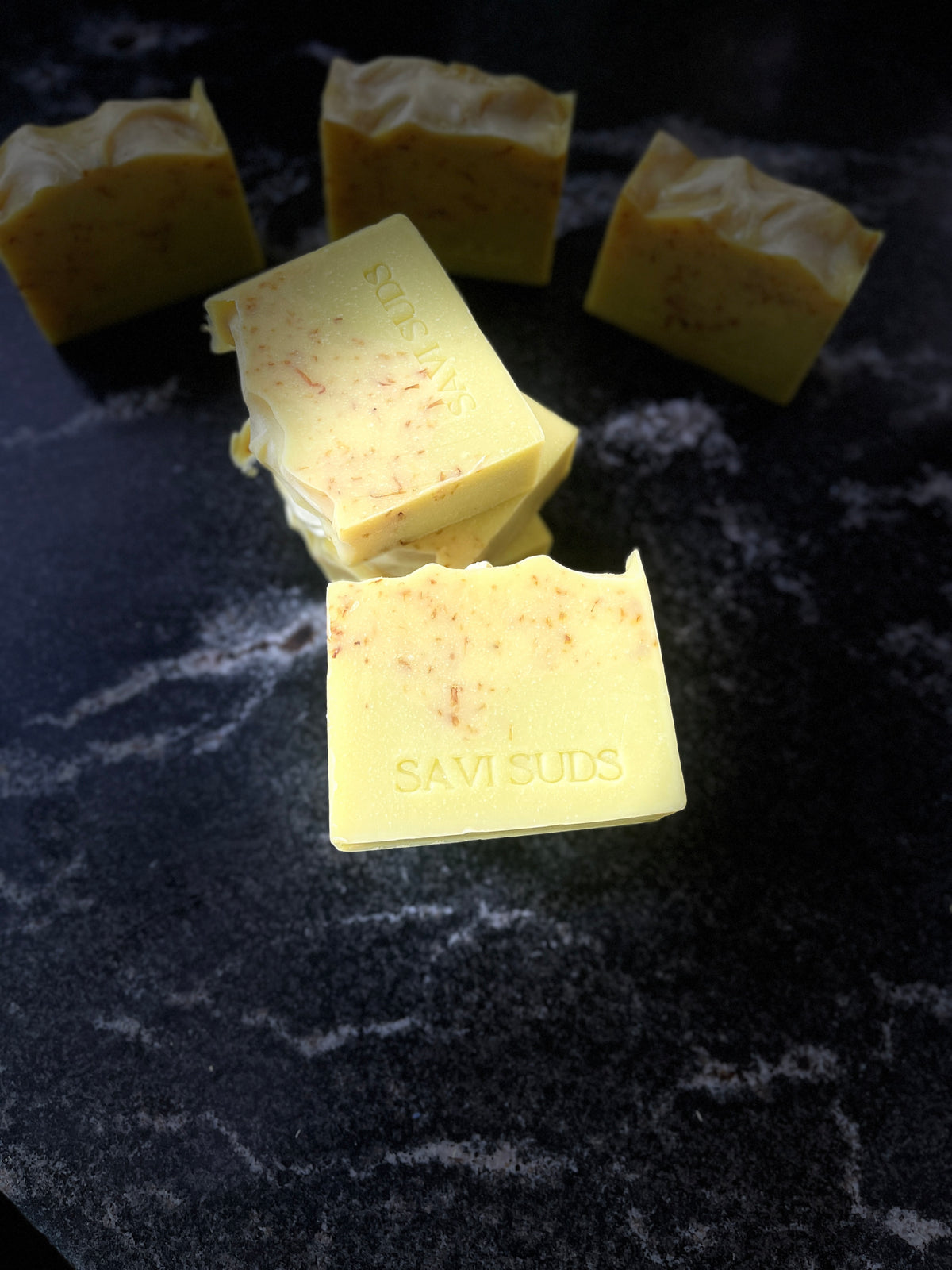 Four square bars of handmade soap, with three in the background and one in the foreground embossed with 'SAVI SUDS', resting on a dark stone surface.