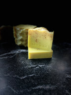 Two square bars of handmade soap stacked on a dark marble surface, with three square bars blurred in the background. The front bar is pale yellow with small botanical speckles, and stamped with the brand name 'SAVISUDS'.