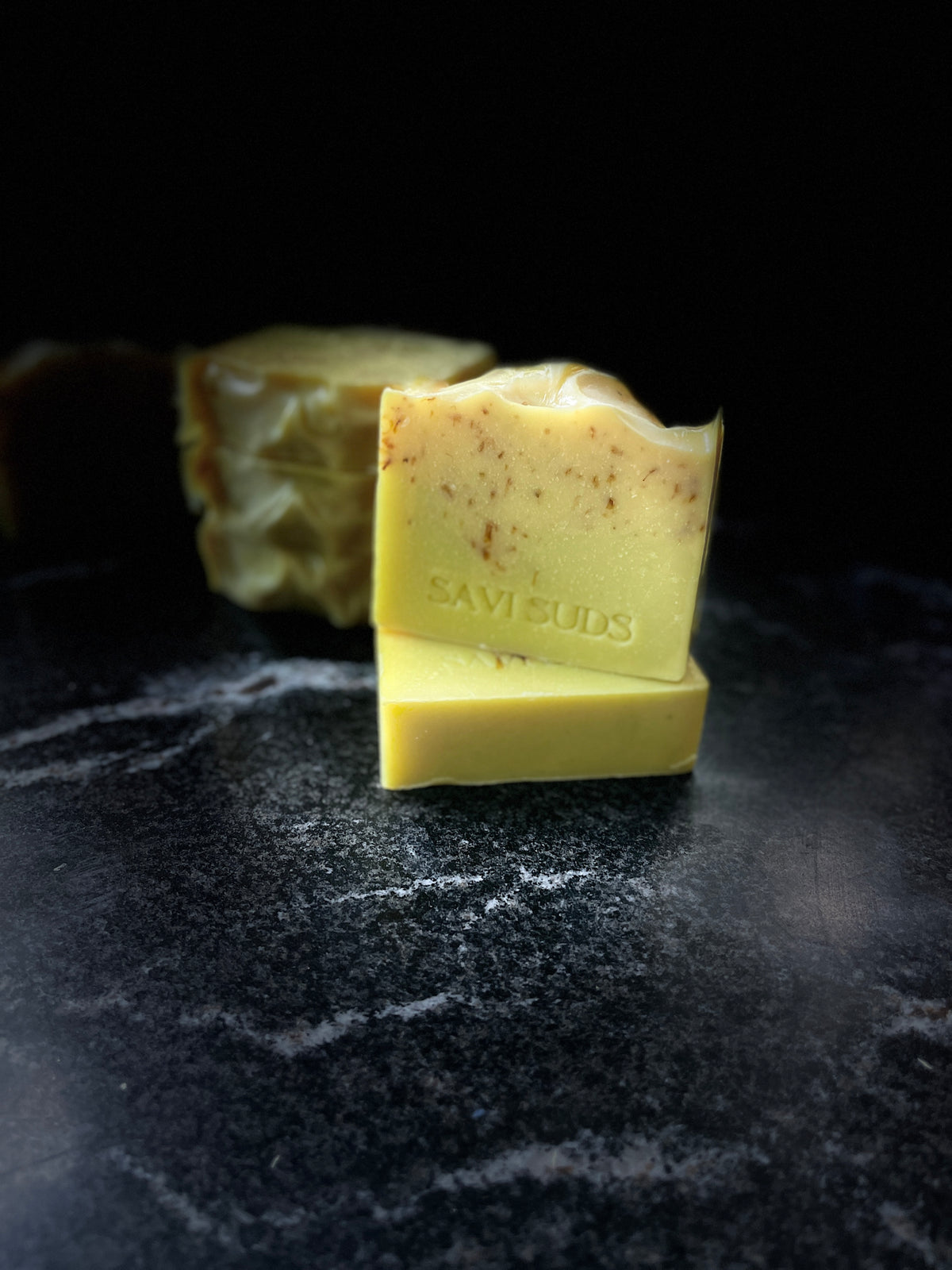 Two square bars of handmade soap stacked on a dark marble surface, with three square bars blurred in the background. The front bar is pale yellow with small botanical speckles, and stamped with the brand name 'SAVISUDS'.