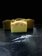Four rectangle bars of handmade soap in a yellow-green hue with botanical inclusions, with one bar in the foreground stamped with the brand name 'SAVI SUDS'. The soap bars are arranged on a dark, textured stone surface against a solid black background.