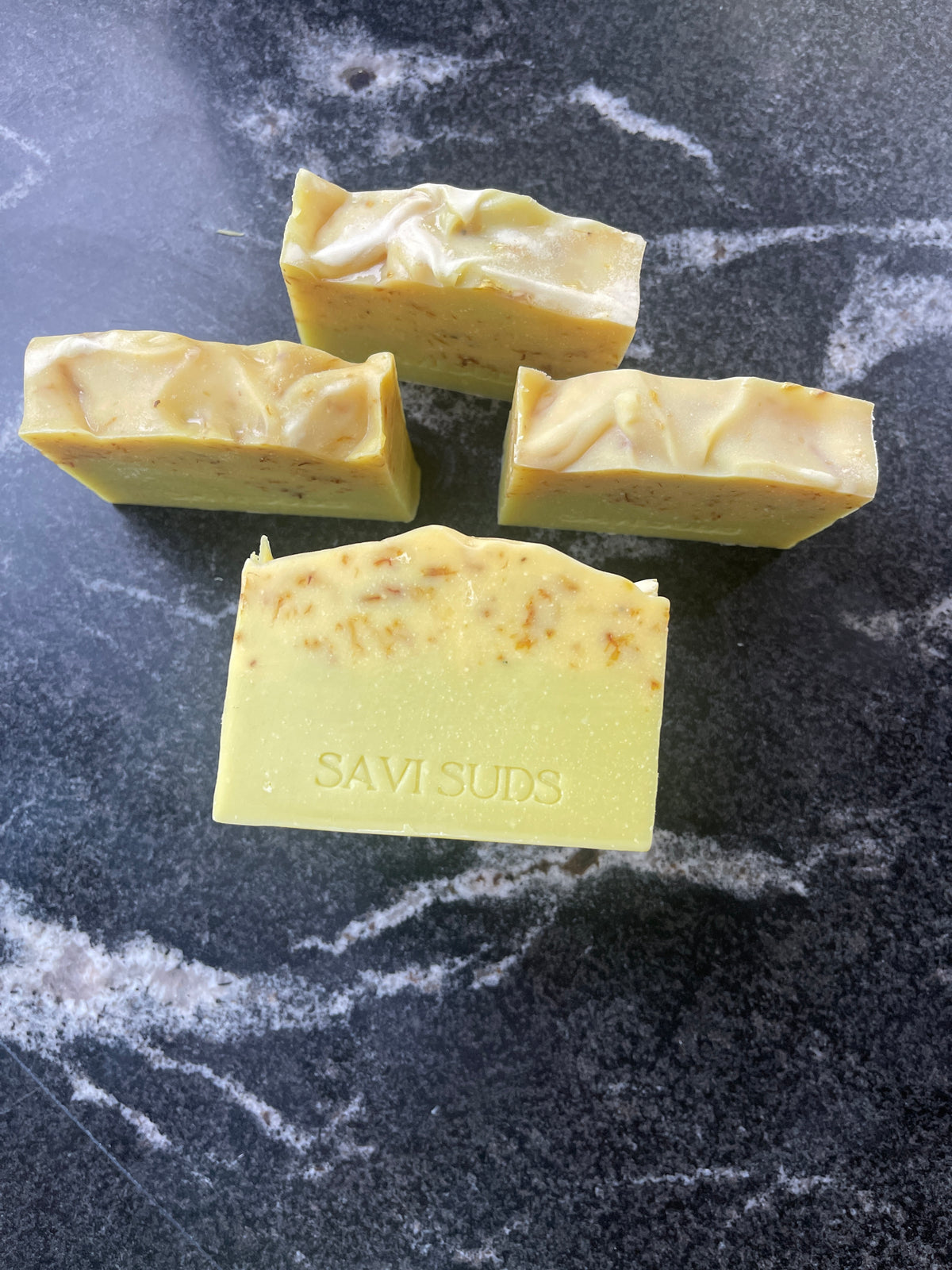 Four rectangle handmade soap bars in a pale yellow-green color with small botanical inclusions, displayed on a dark marble surface. The front bar is stamped with the brand name 'SAVI SUDS'.