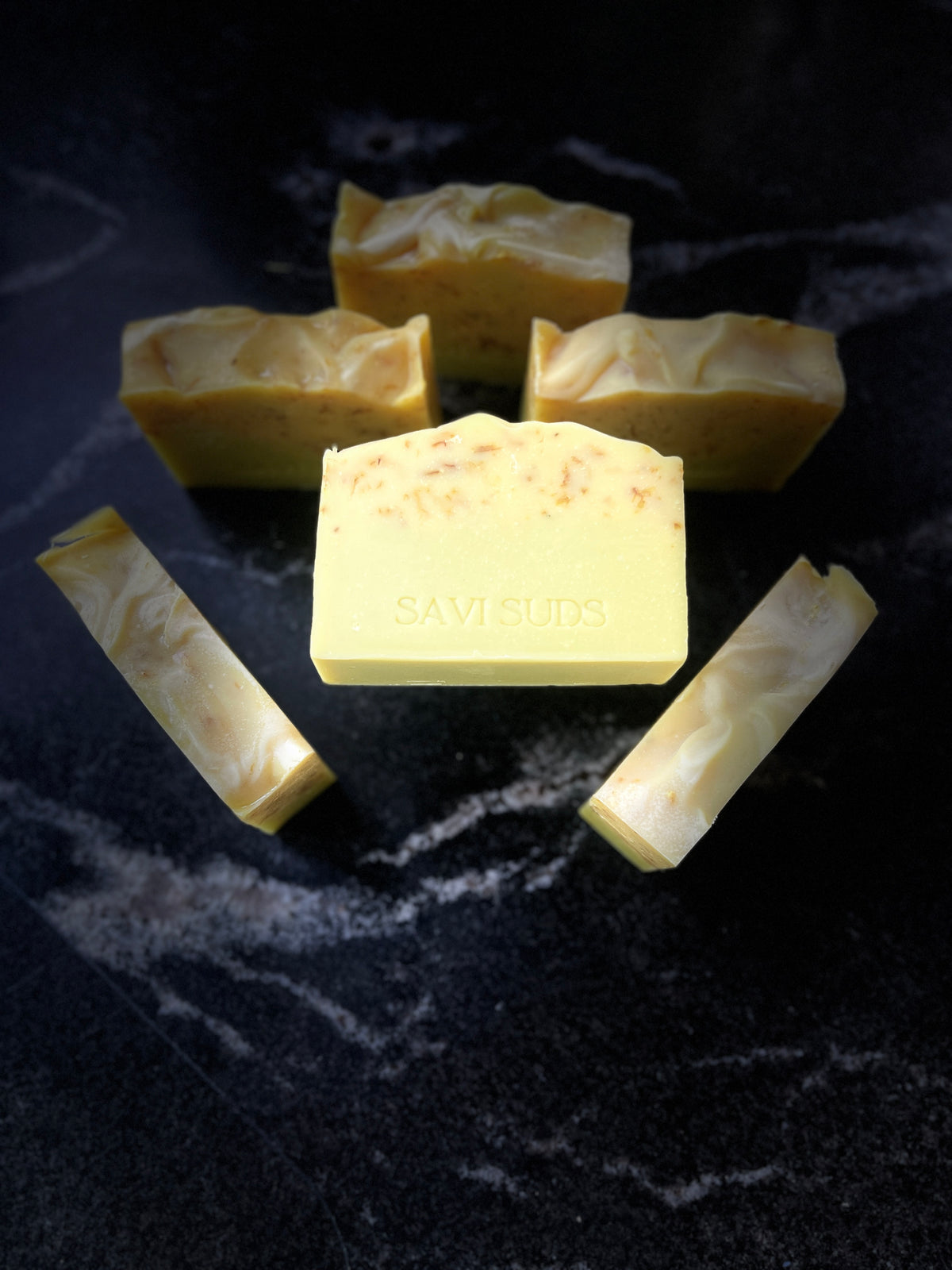 Six rectangle bars of handmade soap in pale yellow and green shades, with one central bar stamped with the brand name 'SAVISUDS', arranged on a dark textured background.
