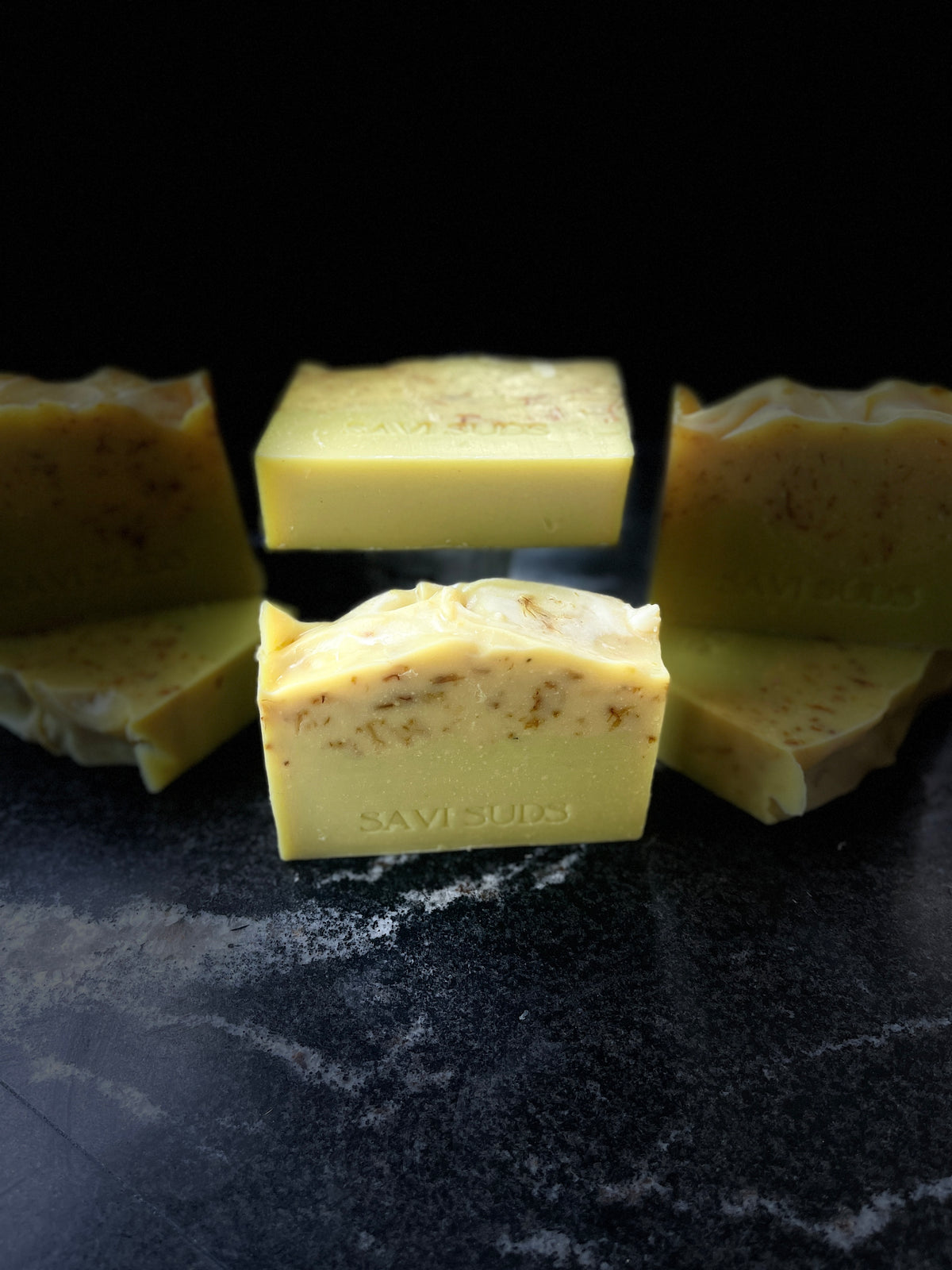 Several rectangle handmade soap bars in a pale yellow tone with embedded botanical pieces, arranged on a dark surface against a black background. The front bar is upright and centered, with other bars partially visible behind it.
