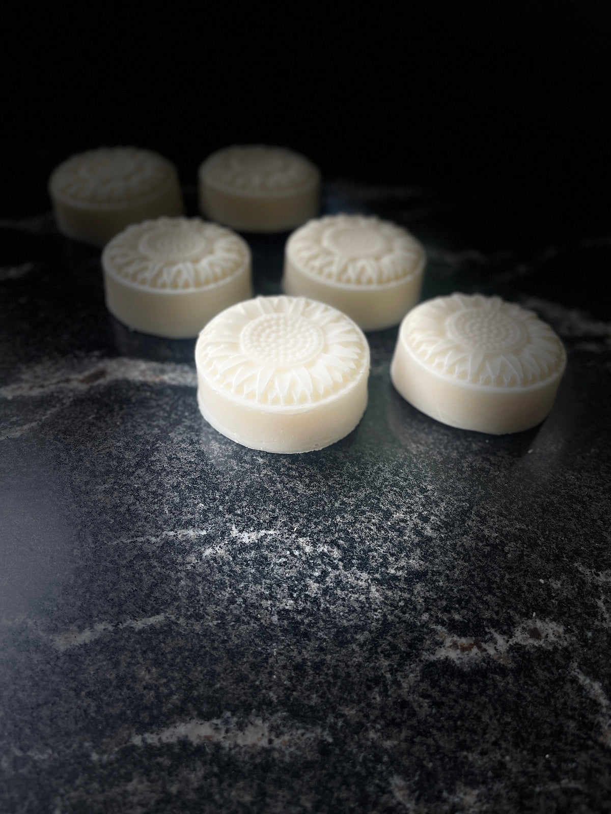 Six round, off-white soap molded into flowers, arranged on a dark, textured stone surface against a black background.