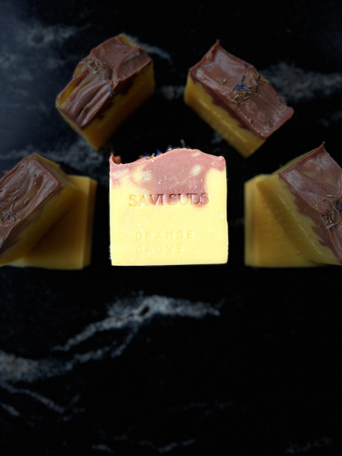 Several artisanal bars of soap arranged on a dark marble surface, with one central bar facing forward that reads 'SAVI SUDS'. The soaps have a pale yellow base with a dark red/brown drop swirl top layer and dried botanicals on some bars.