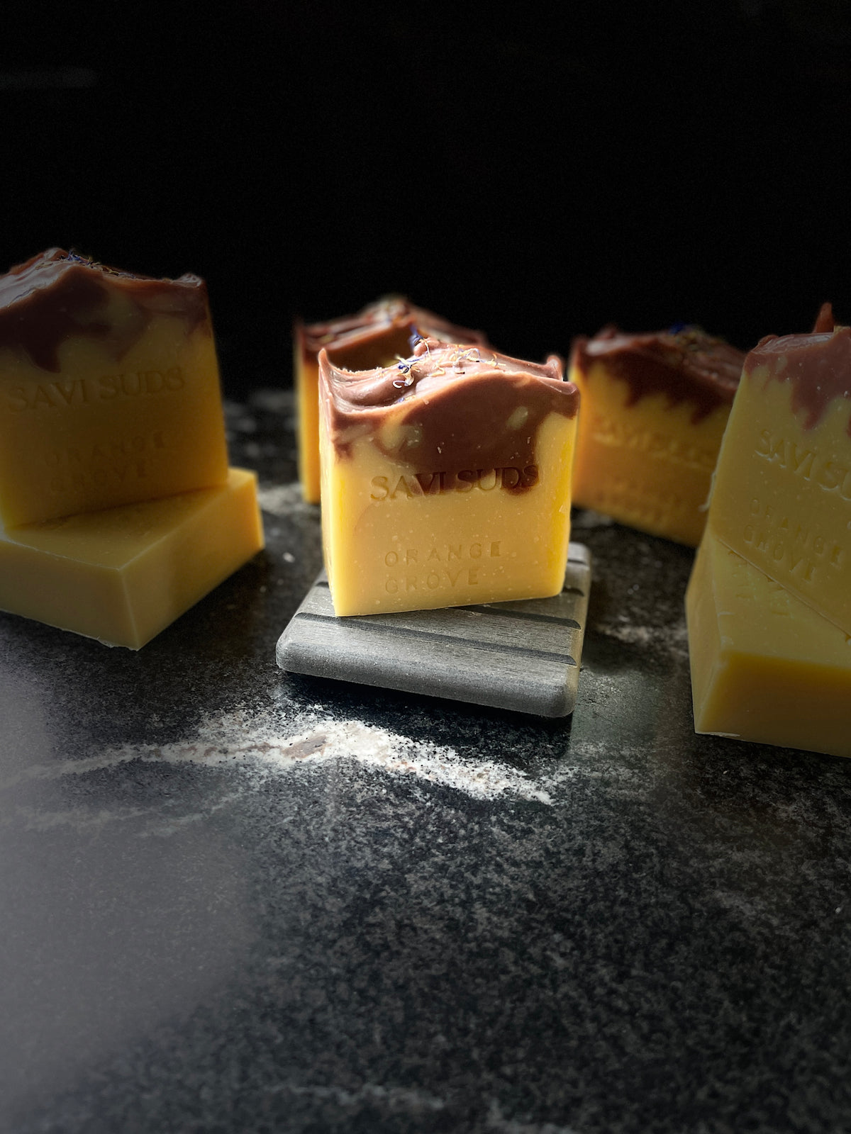 Several square bars of handmade soap in pale yellow and dark red/brown are arranged on a dark surface, with one bar standing upright on a small gray block. The soap has embossed text reading 'Savi Suds' 