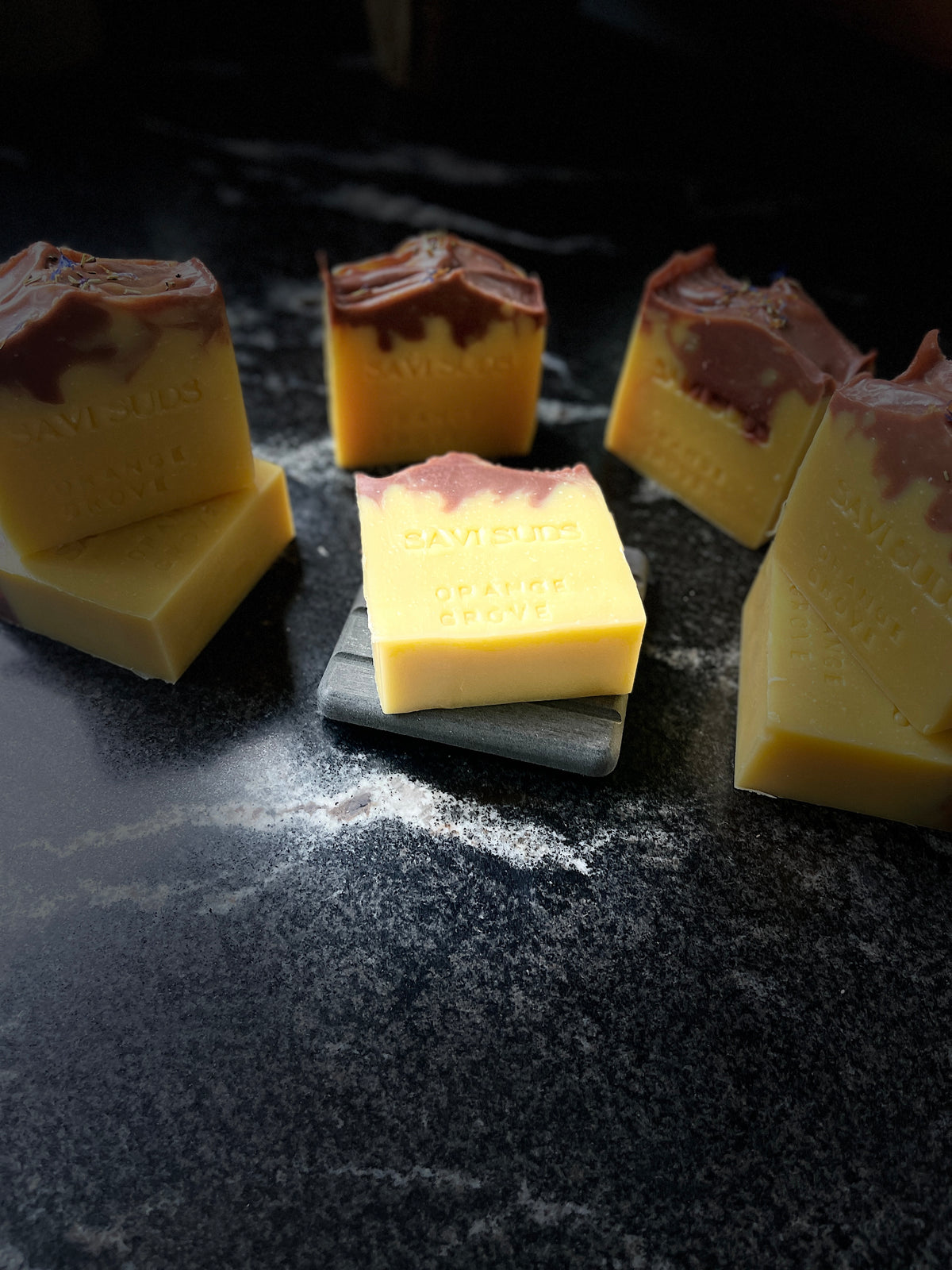 Several handmade soap bars with marbled yellow and dark red/brown tops, arranged on a dark textured surface with one bar resting on a small gray slate tile. The brand name SAVI SUDS is embossed on the soaps.