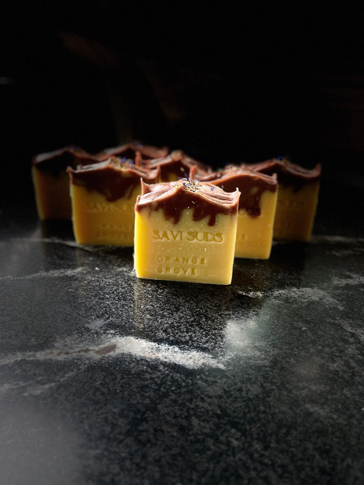 Several square bars of handmade soap in a yellow-orange shade with dark red/brown drop swirl pattern on the top half of the bar. The bars are arranged on a dark marble surface. The front bar is embossed with the brand 'SAVI SUDS'