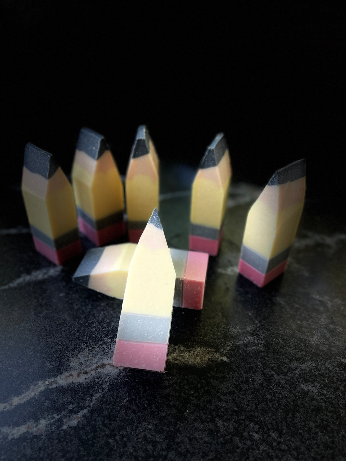 A set of novelty pencil-shaped bath or body soaps standing upright on a dark, textured surface against a black background.