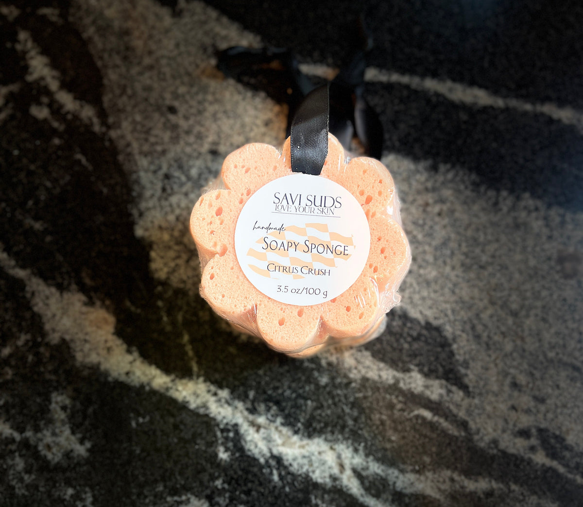 A peach-colored flower-shaped bath sponge by Savi Suds, featuring a white circular label with black text and a black ribbon, resting on a dark marbled surface.