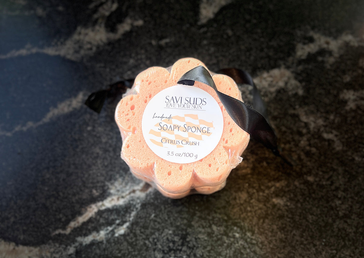 A flower-shaped Savi Suds soap sponge in Citrus scent, wrapped in clear plastic with a black ribbon and a white paper label, placed on a dark marbled surface.
