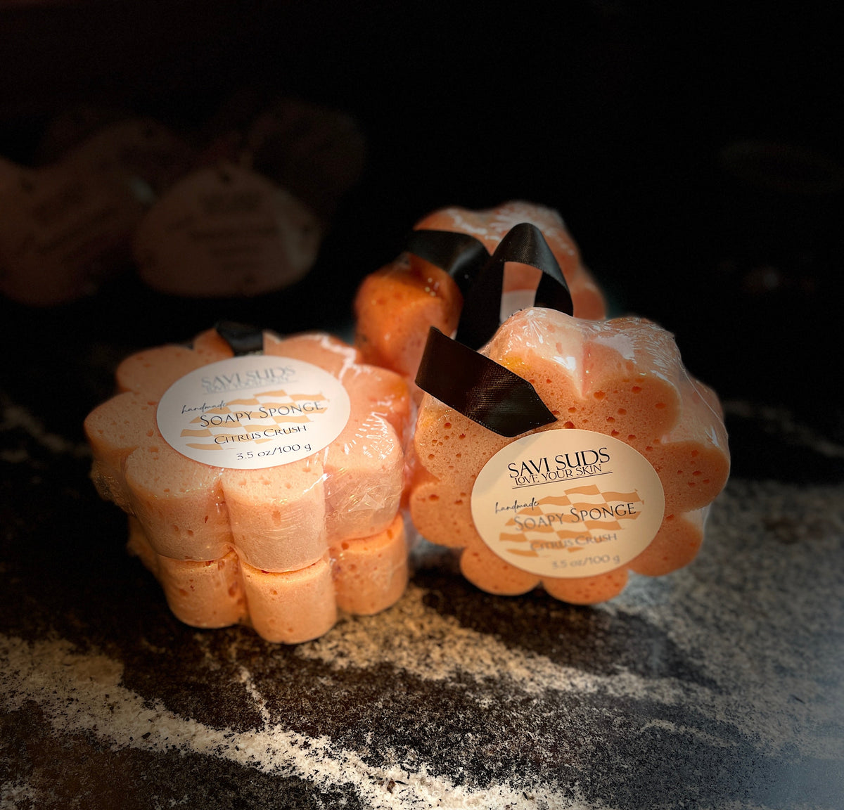 Flower-shaped Savi Suds soap sponges in an orange color, tied with black ribbons and a round white label, set on a dark marble surface 