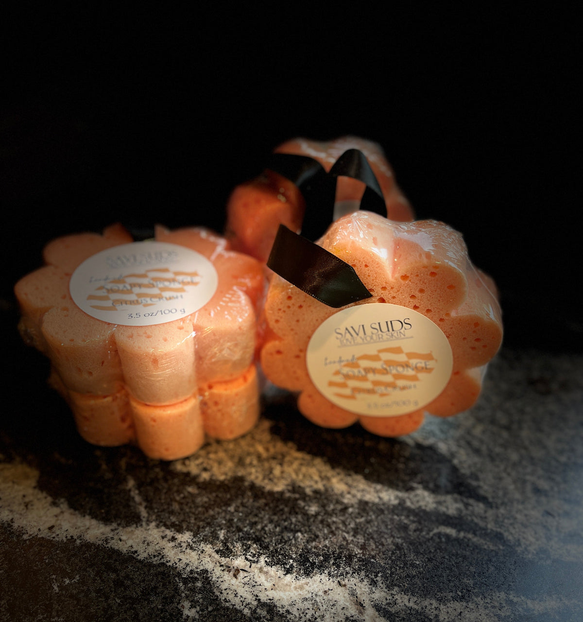 Flower-shaped soap sponges in an orange color packaged in biodegradable plastic with a black ribbon and round white label, set on a dark textured surface.
