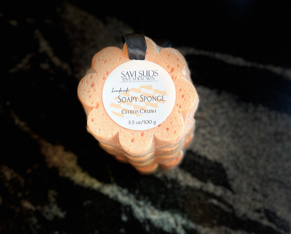 A flower-shaped bath sponge by Savi Suds, Citrus Crush scent, with a white paper label, resting on a dark marbled surface.