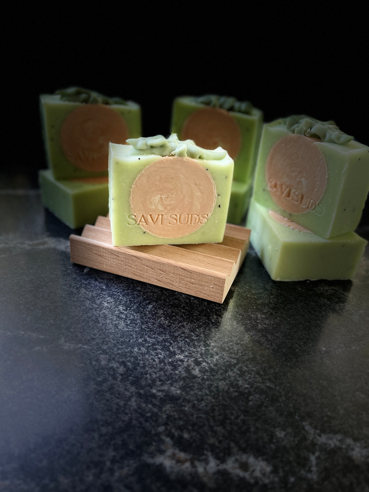 Several handmade square bars of soap in pale green are arranged on a dark surface, with one bar standing upright on a small wooden block in the foreground. Each soap has a round orange swirled center with the brand name SAVI SUDS stamped on the front