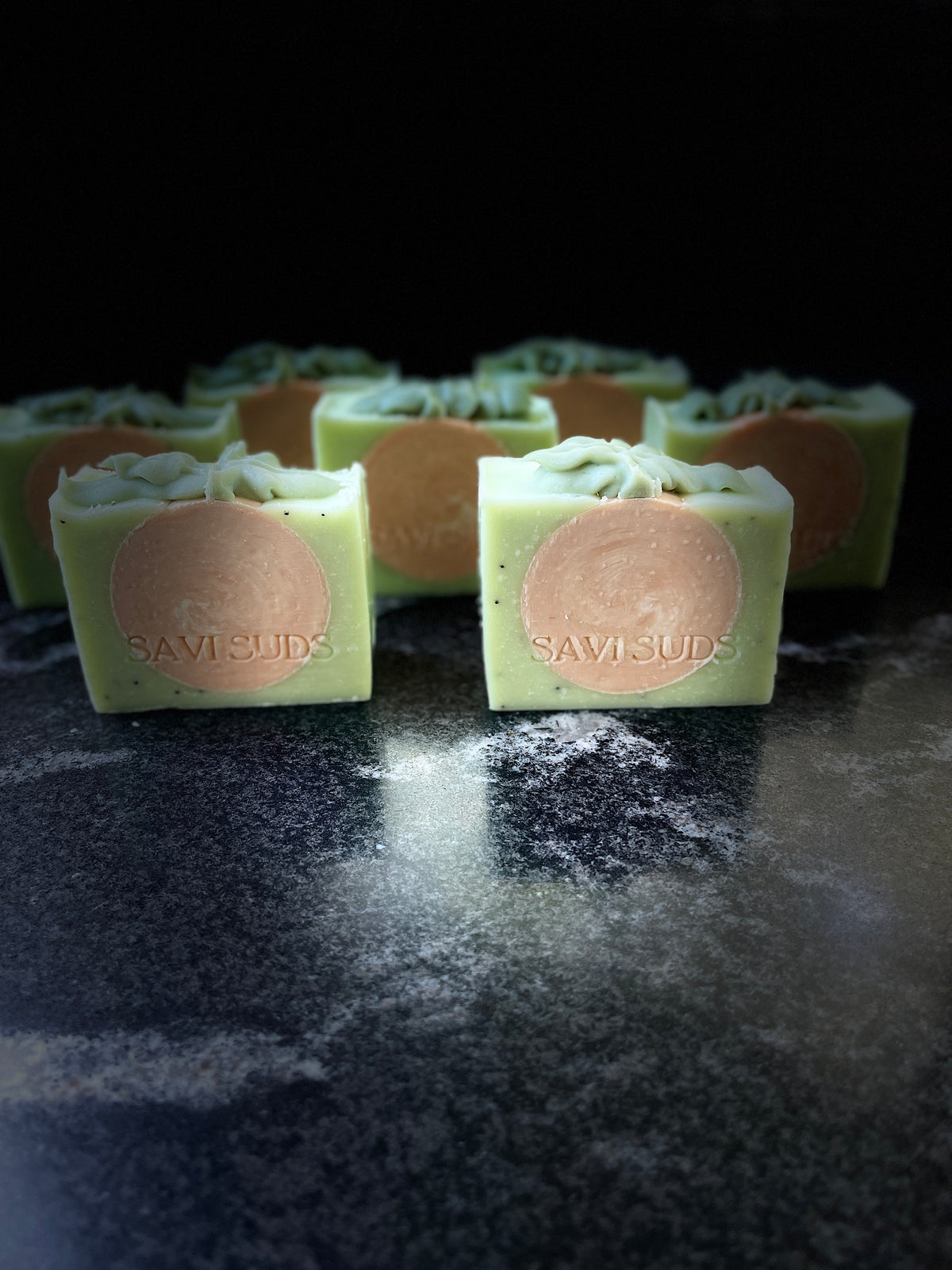 Several square bars of handmade soap in pale green with circular orange swirled centers, arranged on a dark reflective surface against a black background. The front bars show the brand name SAVI SUDS stamped in the center.
