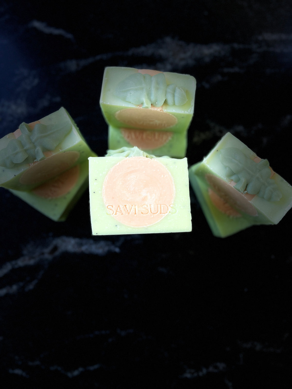Seven square bars of handmade soap in pale green with a circular orange swirl in the middle, arranged on a black marble-like surface. The center bar is upright and shows a stamped brand name SAVI SUDS on the front