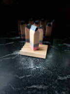 A handmade rectangular pencil-shaped soap with a pink base, gray band, yellow/tan middle, and dark charcoal top, displayed on a small wooden stand against a dark marble surface with more  small pencils blurred in the background.