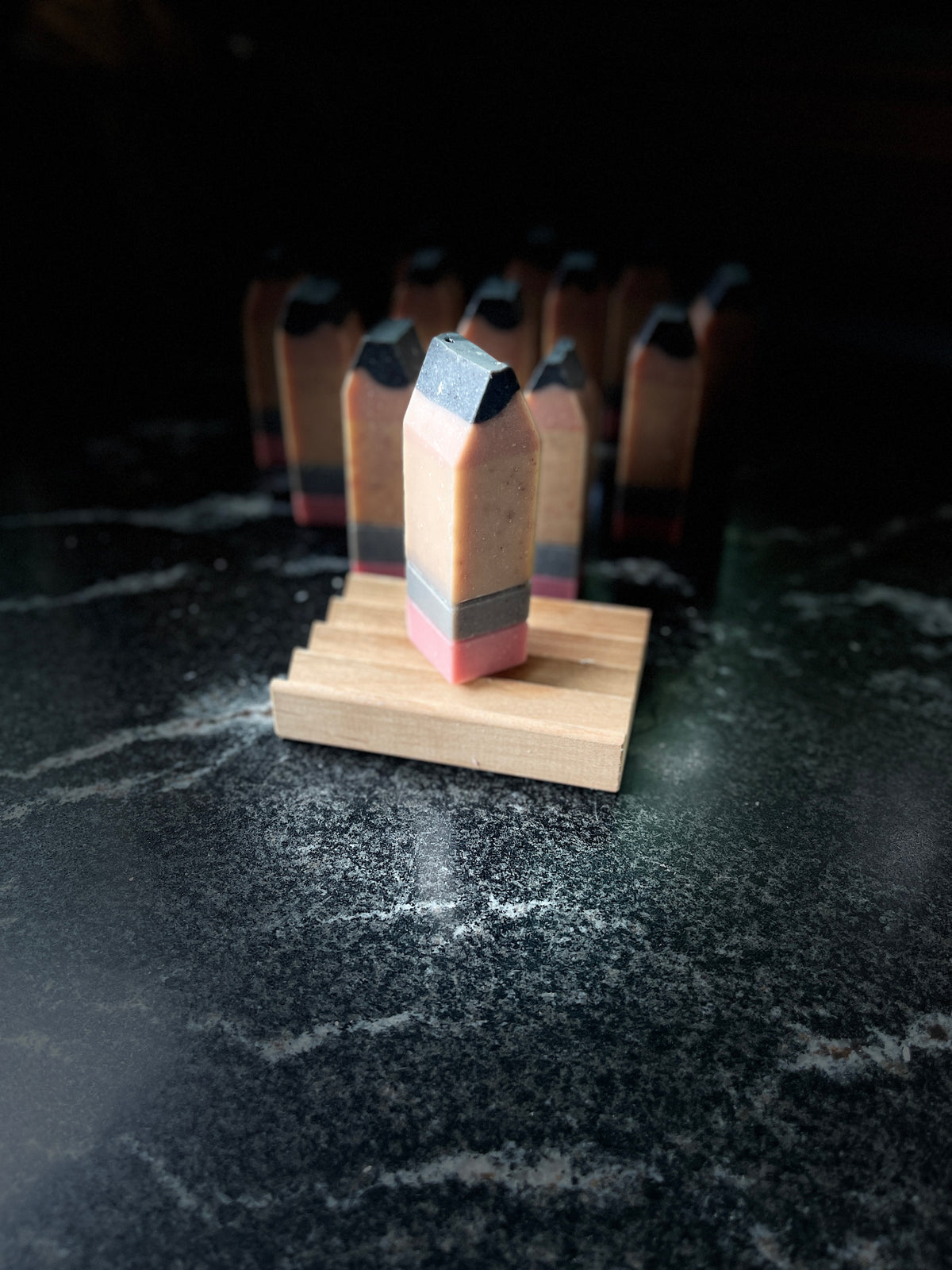 A handmade rectangular pencil-shaped soap with a pink base, gray band, yellow/tan middle, and dark charcoal top, displayed on a small wooden stand against a dark marble surface with more  small pencils blurred in the background.