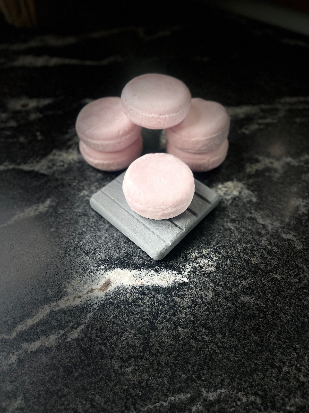 Crystal sands round shampoo bars arranged on a dark marble surface, with one resting on a small gray square dish and the others grouped behind it on a dark marbled surface