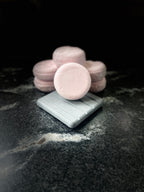 Pink round crystal sands shampoo bars stacked behind a light grey square dish, with one shampoo bar resting on the dish. Displayed on a dark marbled surface