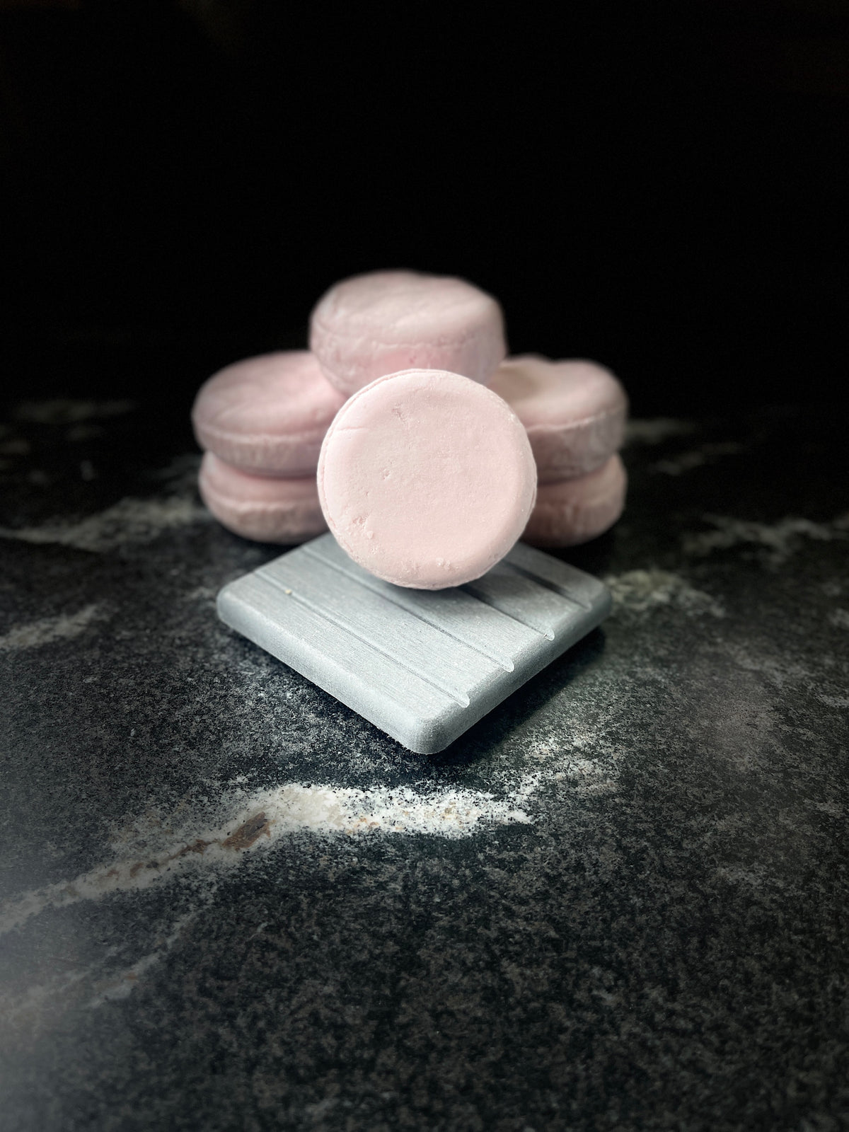 Pink round crystal sands shampoo bars stacked behind a light grey square dish, with one shampoo bar resting on the dish. Displayed on a dark marbled surface