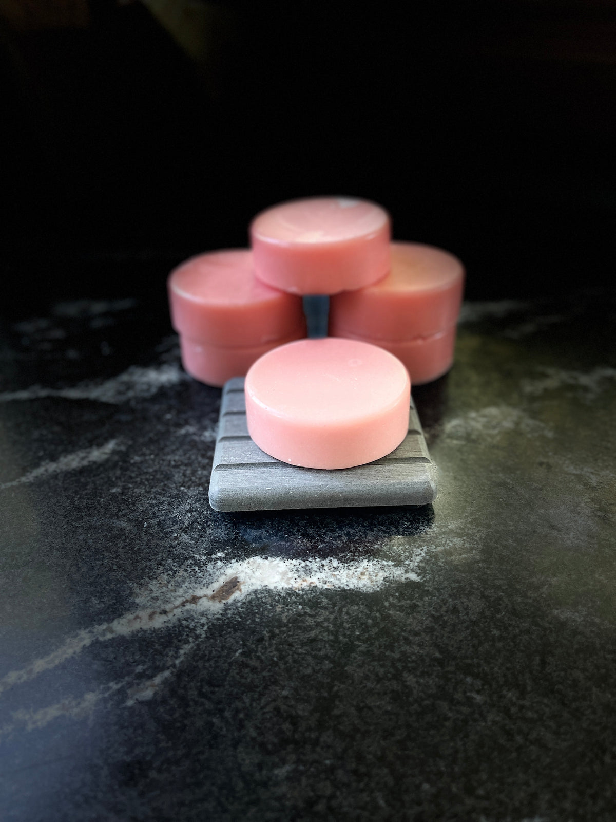 Five pink crystal sands conditioner bars stacked in the background and one resting on a square grey stone in the foreground, set on a dark marble surface
