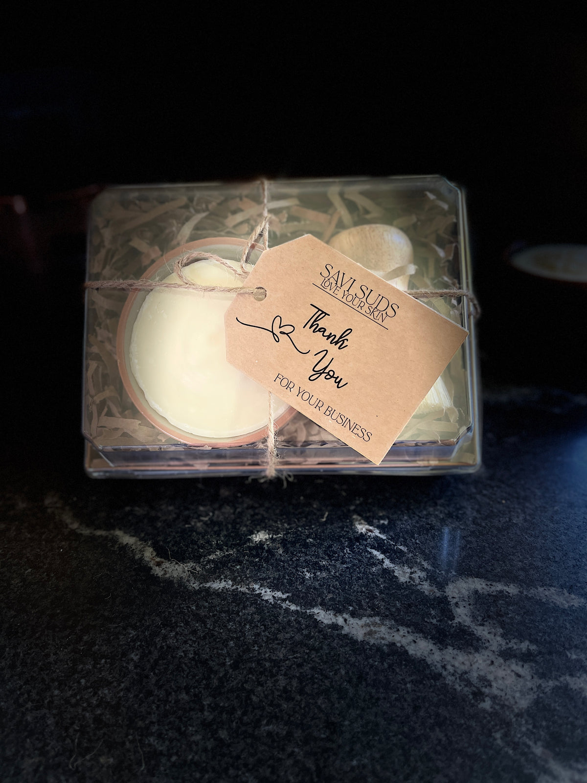 A boxed gift set of solid dish soap in a ramekin and a bamboo dish brush. Packaged in a box with shredded paper around it, and a clear top. A twine string is tied around it with a kraft cardstock tag