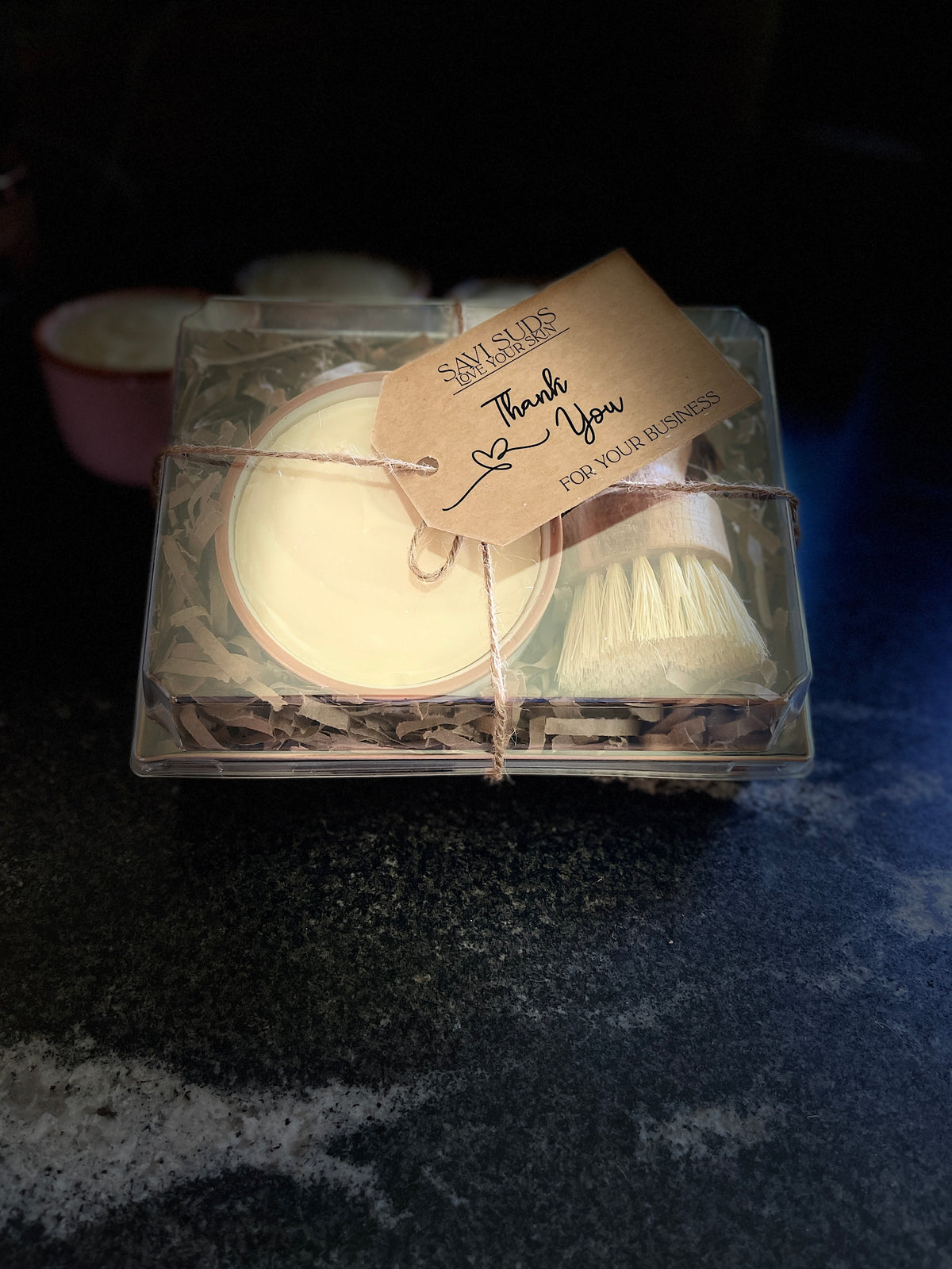 A boxed gift set of solid dish soap in a ramekin and a bamboo dish brush. Packaged in a box with shredded paper around it, and a clear top. A twine string is tied around it with a kraft cardstock tag