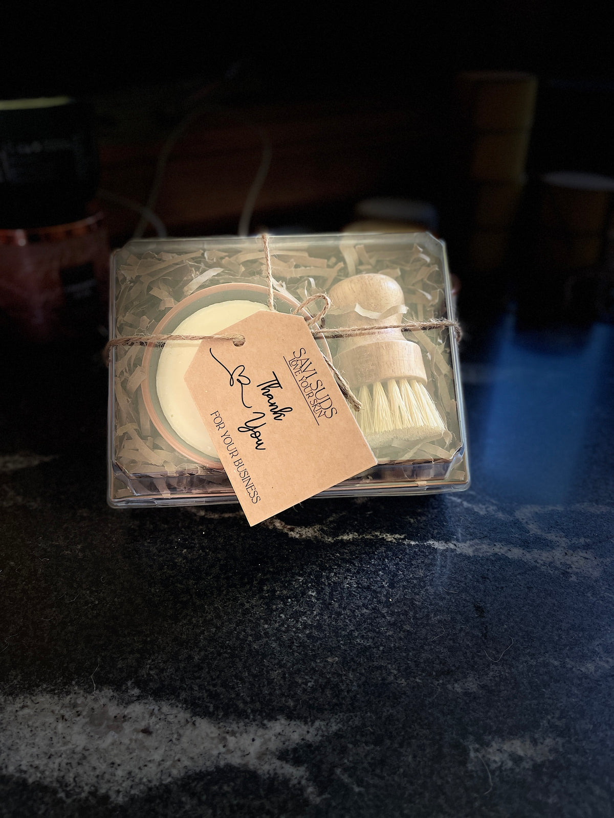 A boxed gift set of solid dish soap in a ramekin and a bamboo dish brush. Packaged in a box with shredded paper around it, and a clear top. A twine string is tied around it with a kraft cardstock tag