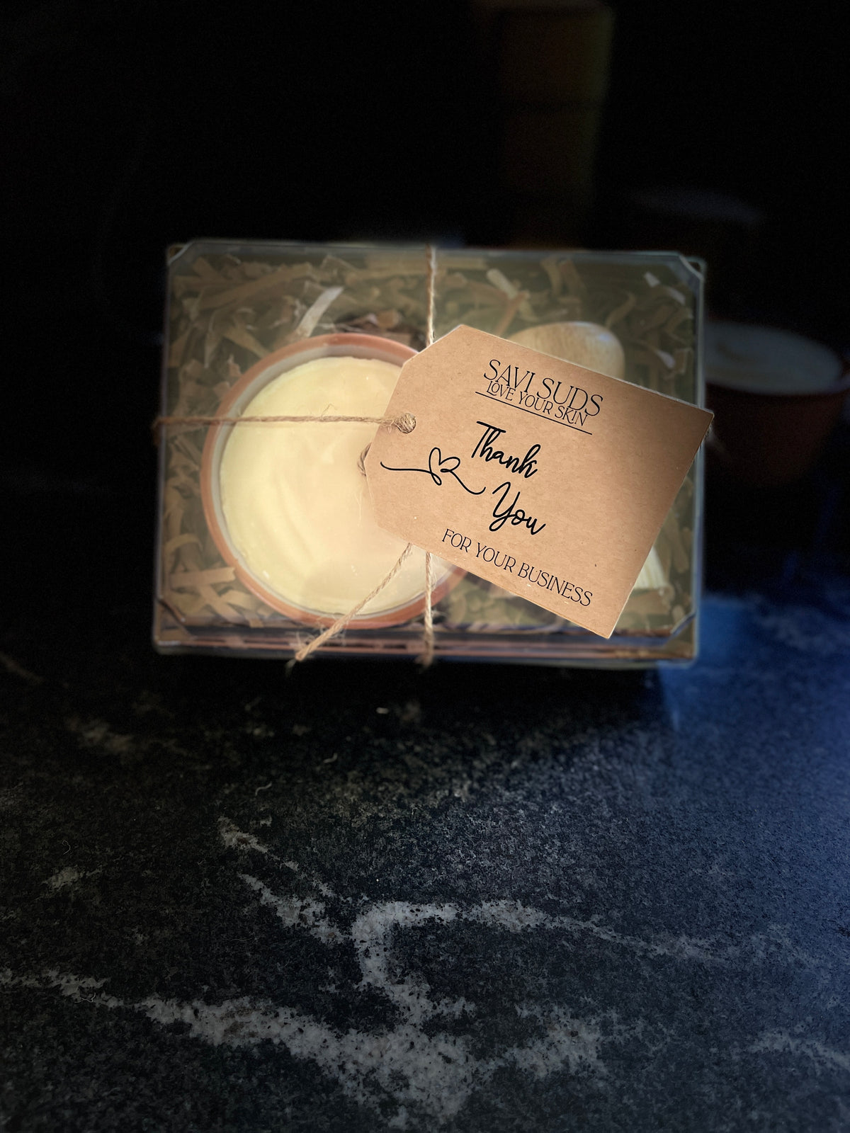 A boxed gift set of solid dish soap in a ramekin and a bamboo dish brush. Packaged in a box with shredded paper around it, and a clear top. A twine string is tied around it with a kraft cardstock tag