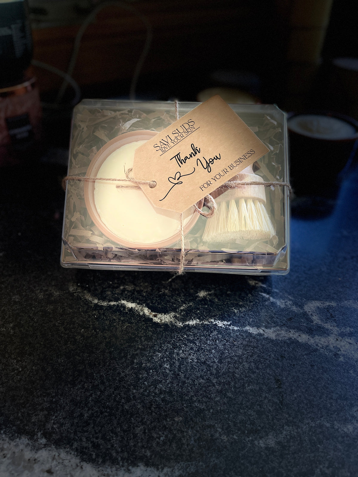 A boxed gift set of solid dish soap in a ramekin and a bamboo dish brush. Packaged in a box with shredded paper around it, and a clear top. A twine string is tied around it with a kraft cardstock tag